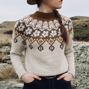 May include: A beige sweater with a brown and cream floral pattern across the yoke and upper chest. The sweater has a brown ribbed collar and cuffs. The person is wearing a hat and has their hands in their pockets.