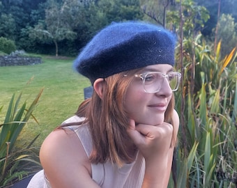 May include: A person wearing a blue beret and clear glasses is looking off to the side. They are wearing a light beige sleeveless shirt and have long brown hair.