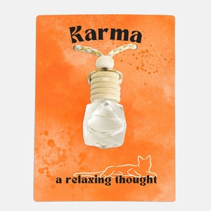 Puede incluir: Un ambientador de coche blanco con una botella de vidrio transparente y una tapa de madera. La botella cuelga de una cuerda y tiene la palabra "Karma" impresa en la tarjeta. La tarjeta también dice "un pensamiento relajante".