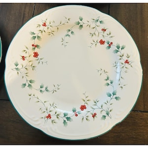 May include: White ceramic plate with a scalloped edge and a green border. The plate is decorated with a holly and berry design in green, red, and white. A second plate is partially visible on the left.