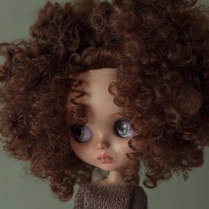 May include: A doll with a pale complexion, large eyes, and a pouty expression. The doll is wearing a brown knitted sweater and has a voluminous head of curly brown hair. The background is a muted green.