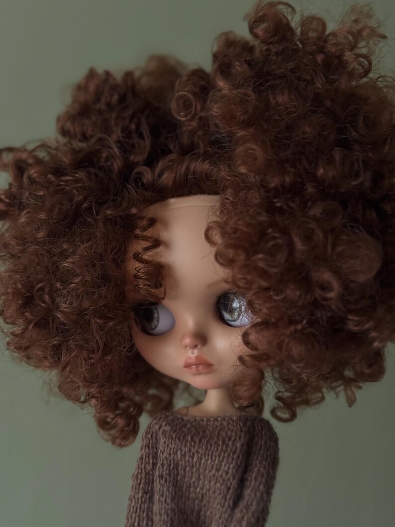 May include: A doll with a pale complexion, large eyes, and a pouty expression. The doll is wearing a brown knitted sweater and has a voluminous head of curly brown hair. The background is a muted green.