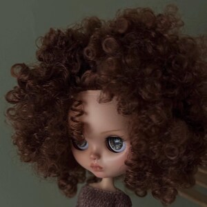 May include: A doll with a detailed face and large, expressive eyes. The doll has a brown curly wig and is wearing a brown sweater. The background is a solid, muted green.