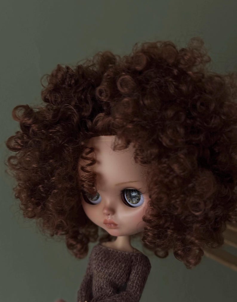 May include: A doll with a detailed face and large, expressive eyes. The doll has a brown curly wig and is wearing a brown sweater. The background is a solid, muted green.