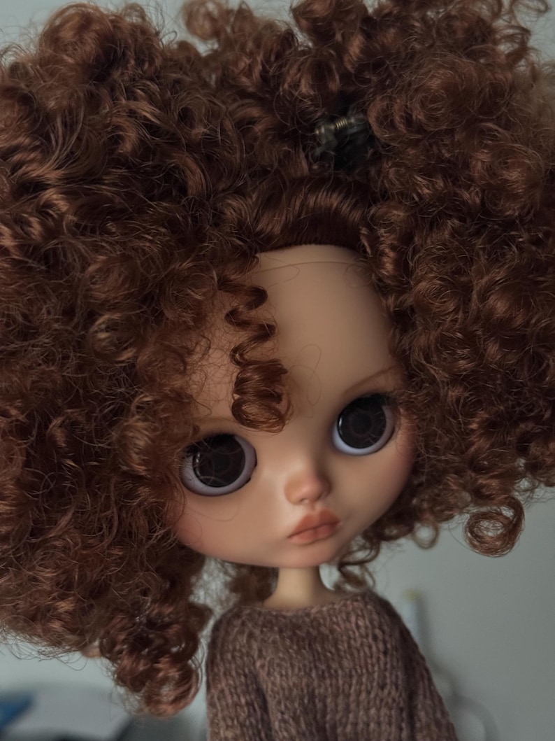 May include: A doll with a fair complexion, large dark eyes, and a neutral expression. The doll has a brown sweater and voluminous, curly auburn hair. A small hair clip is visible in the hair. The doll's face is detailed with subtle makeup.