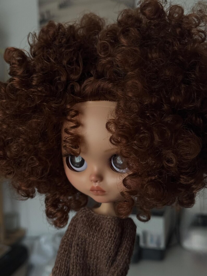 May include: A doll with a fair complexion, large eyes, and a brown curly wig. The doll is wearing a brown jumper. The doll's face has a slight pout, and the eyes are reflective.