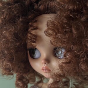 May include: A close-up of a doll's face with large, expressive eyes and full lips. The doll has light skin and a brown sweater. The doll's head is framed by voluminous, curly brown hair.