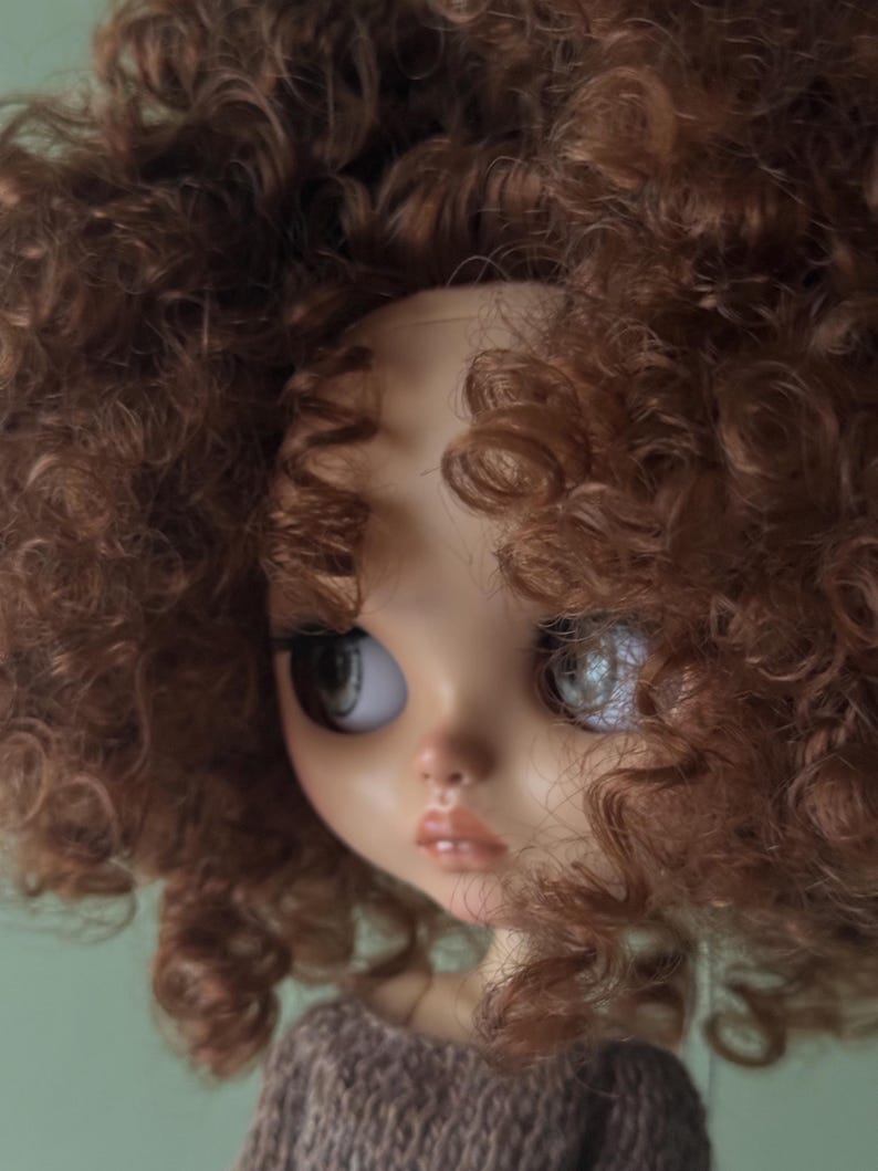 May include: A close-up of a doll's face with large, expressive eyes and full lips. The doll has light skin and a brown sweater. The doll's head is framed by voluminous, curly brown hair.