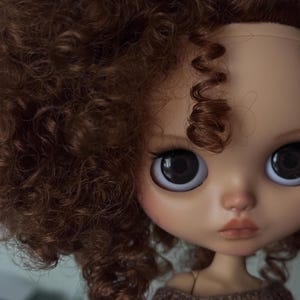 May include: A close-up of a doll's face with large, dark eyes and a neutral expression. The doll has brown, curly hair and is wearing a brown sweater. The doll's skin tone is light with subtle blush.