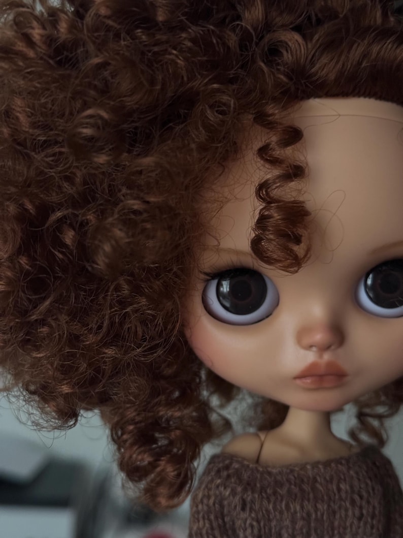 May include: A close-up of a doll's face with large, dark eyes and a neutral expression. The doll has brown, curly hair and is wearing a brown sweater. The doll's skin tone is light with subtle blush.