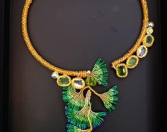 May include: A gold beaded necklace with a pendant of green and gold beaded ginkgo leaves. The necklace is displayed on a black background.