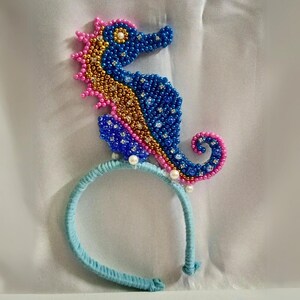 May include: A beaded seahorse headband. The seahorse is blue, gold, and pink, with pearl accents. The headband is light blue and covered in a soft, textured material. A whimsical accessory.