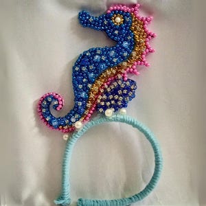 May include: A beaded seahorse headband in blue, gold, and pink. The seahorse is embellished with small beads and rhinestones, and is attached to a light blue headband. The seahorse is approximately 4 inches tall.