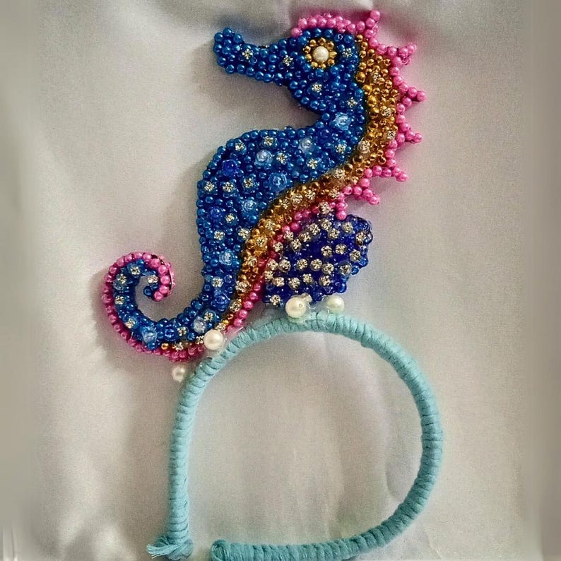 May include: A beaded seahorse headband in blue, gold, and pink. The seahorse is embellished with small beads and rhinestones, and is attached to a light blue headband. The seahorse is approximately 4 inches tall.