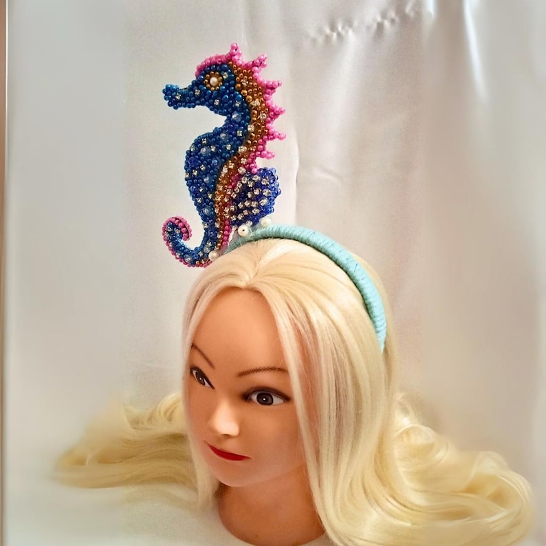 May include: A beaded seahorse headpiece in blue, pink, and gold, mounted on a light blue headband. The seahorse is intricately detailed with small beads and pearls. The headpiece is displayed on a mannequin head with blonde hair.