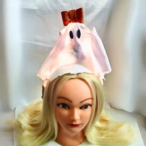 May include: A Halloween headpiece in the shape of a ghost. The headpiece is made of light pink fabric with two black oval eyes. A small orange glitter bow is attached to the top. The headpiece is displayed on a mannequin head with blonde hair.