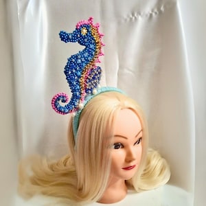 May include: A beaded seahorse headband in blue, pink, and gold. The seahorse is embellished with small beads and sits atop a light blue headband adorned with pearls. The accessory is displayed on a mannequin head with blonde hair.