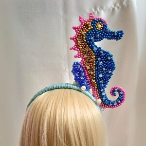 May include: A beaded seahorse headband. The seahorse is blue, pink, and gold, with pearl accents. The headband is light blue. The seahorse is the focal point, with a detailed, textured design.