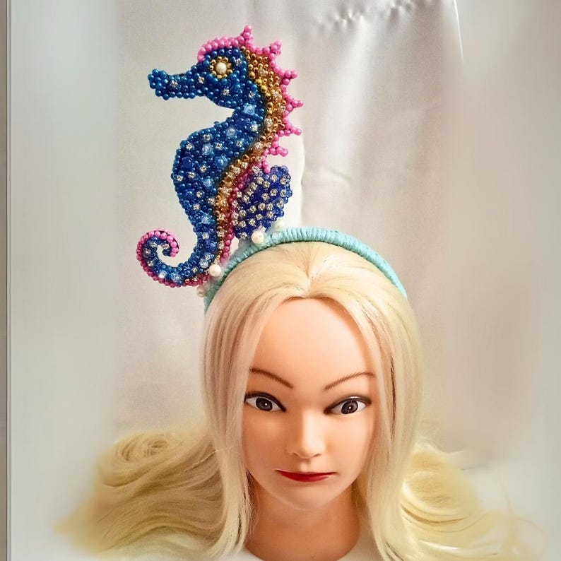 May include: A blue, pink, and gold beaded seahorse headpiece. The seahorse is attached to a light blue headband. The headpiece is on a mannequin head with blonde hair and red lipstick.
