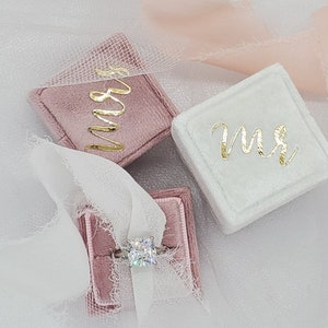 May include: Three velvet ring boxes, two are pink and one is white. The pink boxes have the word "Mrs." written in gold script. The white box has the word "Mr." written in gold script. A diamond ring is in one of the pink boxes.