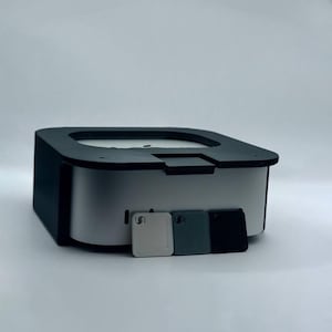Puede incluir: Una caja negra y plateada con una abertura cuadrada en la parte superior. Hay tres pequeñas baldosas cuadradas delante de la caja, una blanca, una gris y una negra.