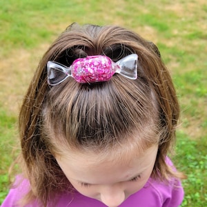 May include: A clear, candy-shaped hair clip with a pink glitter center is secured in a child's hair. The hair clip is made of clear plastic and has a bow-like shape on each end. The child is wearing a purple shirt.