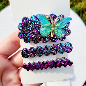 May include: Set of three sparkly hair clips featuring a purple and blue glitter finish. The largest clip has a turquoise butterfly charm.