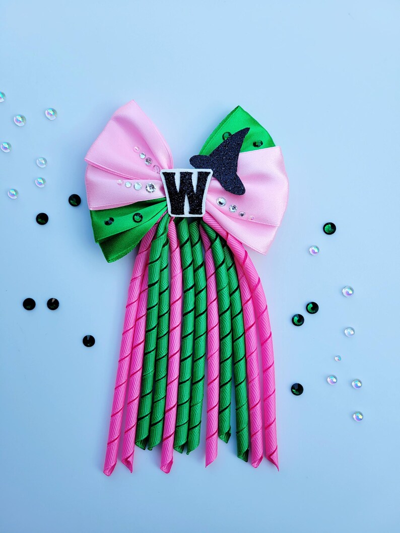 May include: A pink and green hair bow with a black glitter witch hat and a white letter "W" in the center. The bow has pink and green ribbons with a spiral design, and is decorated with clear and black sequins.