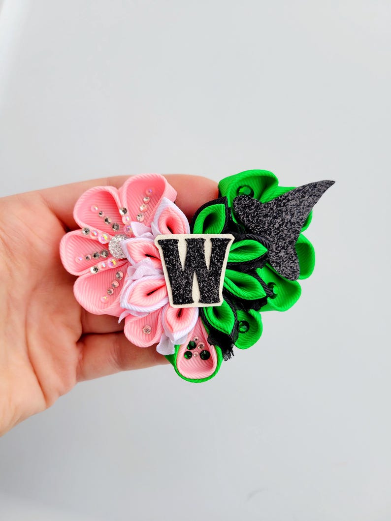 May include: A handmade hair clip featuring a floral design with pink and white petals, green leaves, and a black and white letter "W". The clip is embellished with rhinestones and small green beads. The overall design is a combination of floral and geometric elements.
