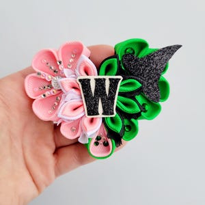 May include: A handmade hair accessory featuring a pink and white flower design with rhinestone accents, and a green and black section. A black and white letter "W" is in the center. The accessory is held in a hand.