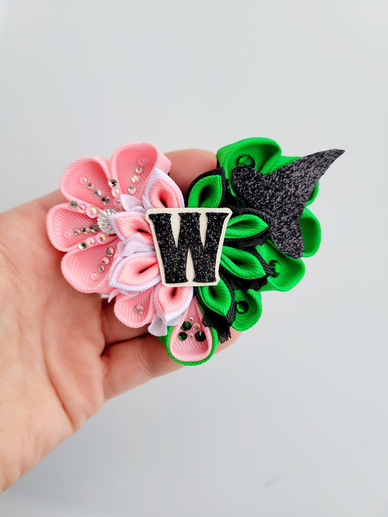 May include: A handmade hair accessory featuring a pink and white flower design with rhinestone accents, and a green and black section. A black and white letter "W" is in the center. The accessory is held in a hand.
