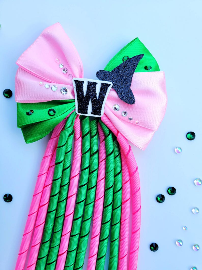 May include: A pink and green hair bow with a black glitter accent shaped like a bird. The bow features the letter "W" and is embellished with rhinestones. The bow has long, spiraled pink and green streamers.