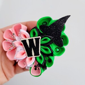 May include: A handmade hair accessory featuring a cluster of fabric flowers in pink and green, accented with a black glittery witch hat and a white letter "W". The accessory is embellished with small rhinestones.