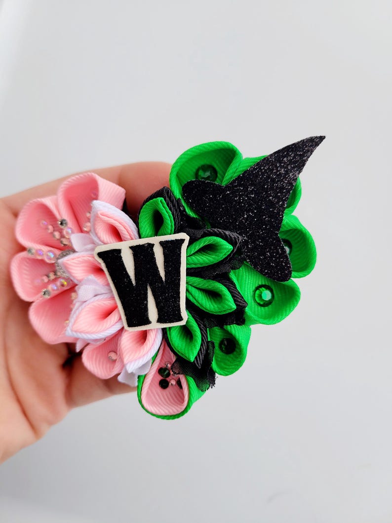 May include: A handmade hair accessory featuring a cluster of fabric flowers in pink and green, accented with a black glittery witch hat and a white letter "W". The accessory is embellished with small rhinestones.