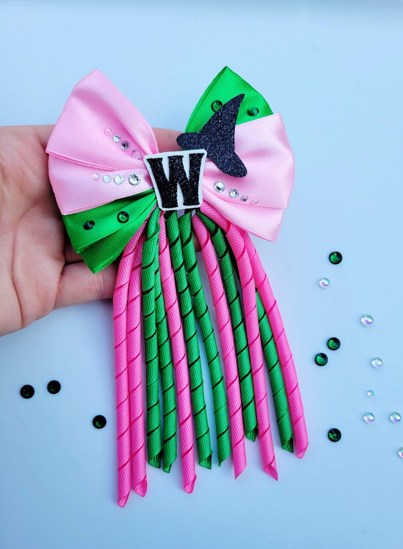 May include: A pink and green hair bow with a black glitter witch hat and the letter "W". The bow features a pink satin bow, green accents, and pink and green spiral ribbons. Rhinestones are scattered around the bow.
