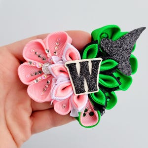May include: A handmade hair clip featuring a pink and green floral design. The pink flower has rhinestone accents and a white center. The green flower has a black accent and a black and white letter "W".