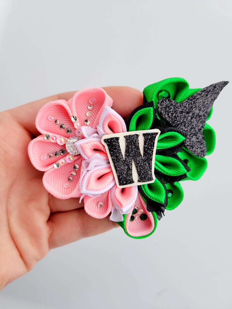 May include: A handmade hair clip featuring a pink and green floral design. The pink flower has rhinestone accents and a white center. The green flower has a black accent and a black and white letter "W".