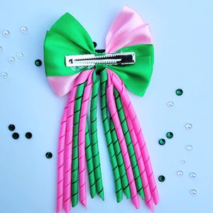 May include: A vibrant hair bow featuring a green and pink color scheme. The bow has a large, layered design with a metal clip for secure attachment. The bow's streamers are a mix of twisted green and pink ribbons.
