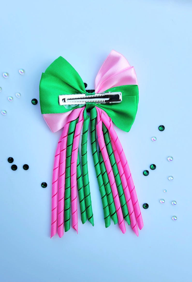 May include: A vibrant hair bow featuring a green and pink color scheme. The bow has a large, layered design with a metal clip for secure attachment. The bow's streamers are a mix of twisted green and pink ribbons.