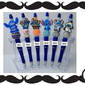 May include: Six blue pens with different Stitch character charms. The charms include Stitch holding a pumpkin, Stitch wearing a cowboy hat, Stitch with a lei, Stitch wearing a Santa hat, Stitch wearing a cow costume, and Stitch wearing a snowman costume. The pens are on a clear plastic stand.