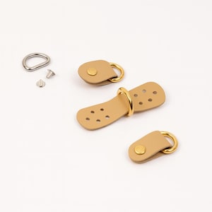 May include: A set of tan leather bag hardware with gold-coloured metal rings and rivets. Includes two small pieces, one larger piece, a metal ring, and screws. Ideal for leathercraft projects.