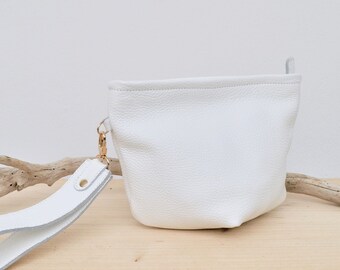 Può includere: Una pochette in pelle bianca con una tracolla bianca e dettagli dorati. La pochette è appoggiata su una superficie in legno.
