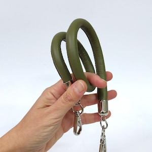 Può includere: Un cinturino in pelle verde con fibbie argentate. Il cinturino è ad anello e viene tenuto in una mano.