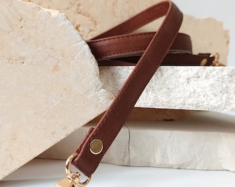 May include: A brown leather strap with a gold metal clasp. The strap is folded and placed on a white surface.