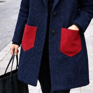 Può includere: Un cappotto blu navy con due grandi tasche rosse. Il cappotto ha un classico colletto a revers e due bottoni neri. La persona porta una borsa tote nera. Il cappotto è realizzato in un tessuto strutturato.