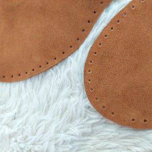 May include: Close-up of two brown suede leather bag bases with pre-punched holes for stitching. The bases are oval-shaped and rest on a white, fluffy surface. The leather has a soft, textured appearance.