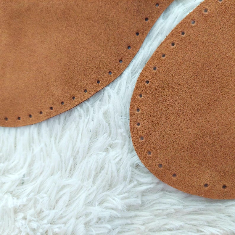 May include: Close-up of two brown suede leather bag bases with pre-punched holes for stitching. The bases are oval-shaped and rest on a white, fluffy surface. The leather has a soft, textured appearance.