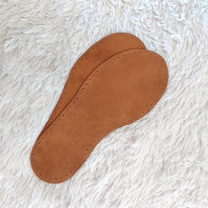 May include: Two brown leather insoles with a series of small holes around the edges. The insoles are oval-shaped and designed to fit inside shoes. The insoles are made of a soft, suede-like material.
