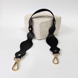 Könnte beinhalten: Schwarzer Leder-Taschenriemen mit welligem Design und goldenen Beschlägen. Der Riemen hat zwei goldene Verschlüsse.