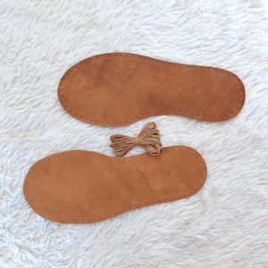 May include: Two brown leather shoe soles with pre-punched holes for stitching, and a bundle of brown twine. The soles are oval-shaped, and the twine is tied in a bow. The items are on a white, fluffy surface.